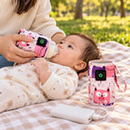 Bébé buvant son biberon sur un plaid de pique-nique, à côté du chauffe-biberon de voyage branché sur une batterie externe